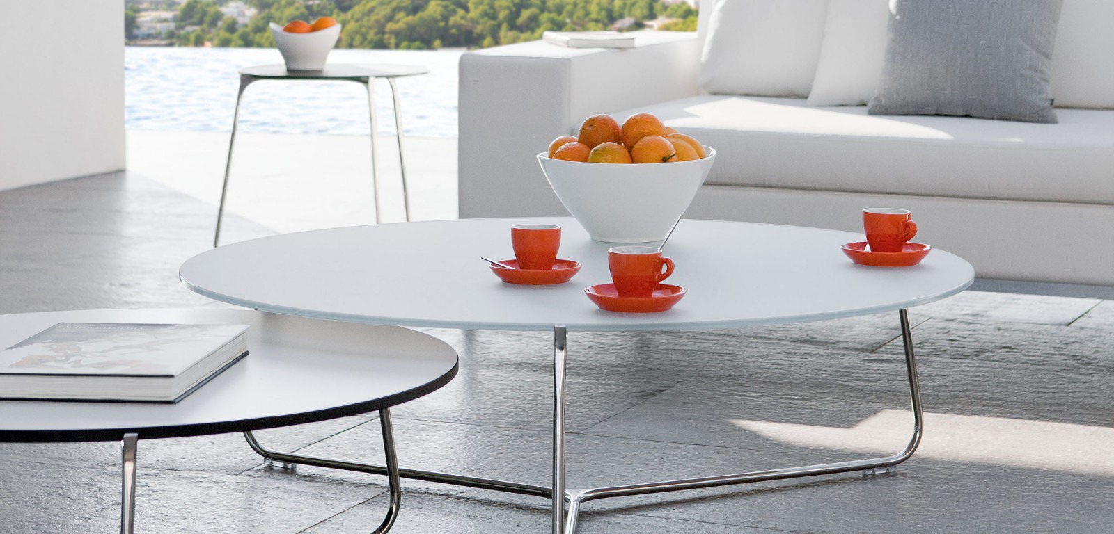 Mood Contemporary Coffee Table — Modern living room with a white sofa, oval coffee table, red cups, and oranges, overlooking the water