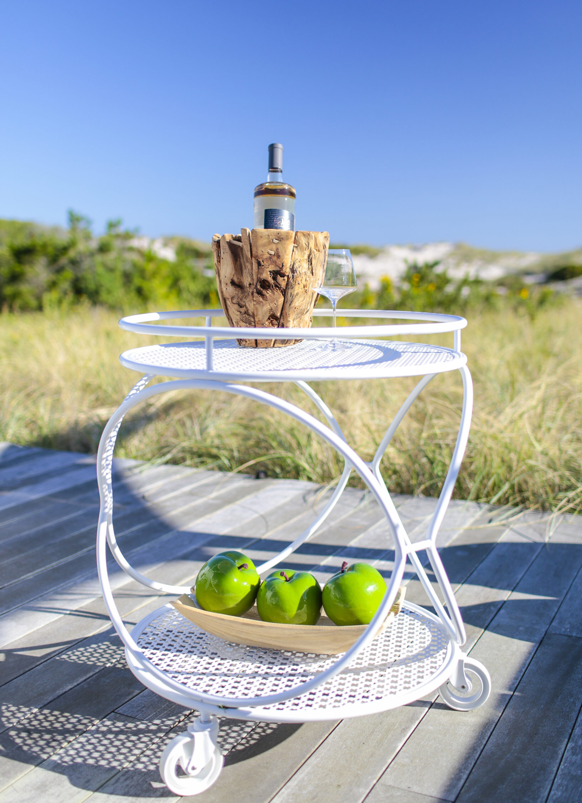 White serving cart with wine bottle and green apples outdoors.