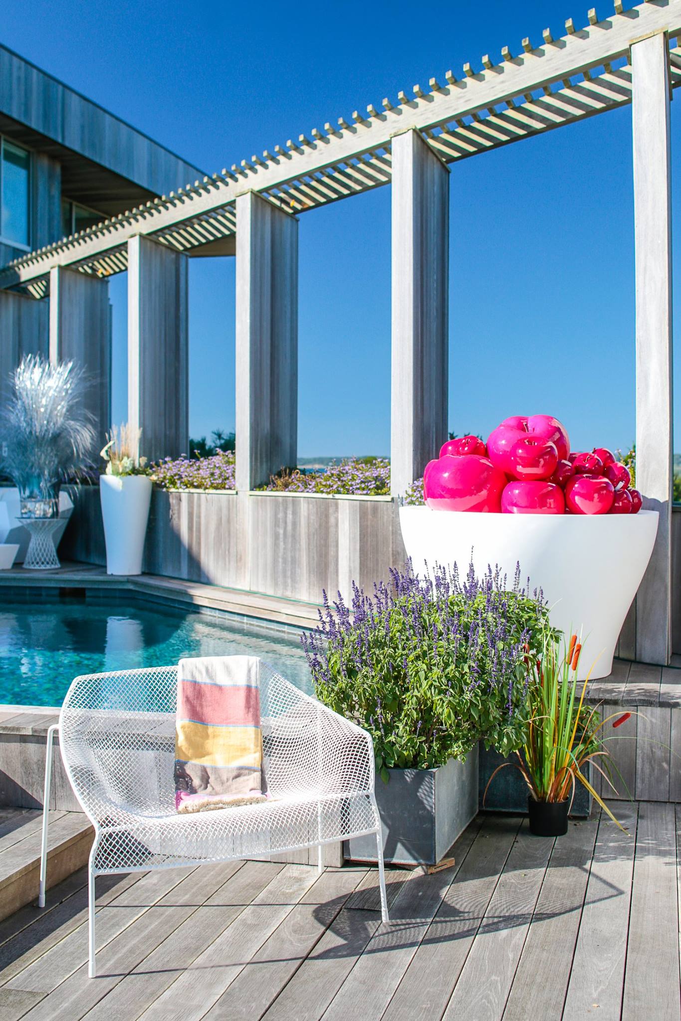 Couture Outdoor poolside area with a white mesh chair, potted plants, and a bowl of shiny pink apples.