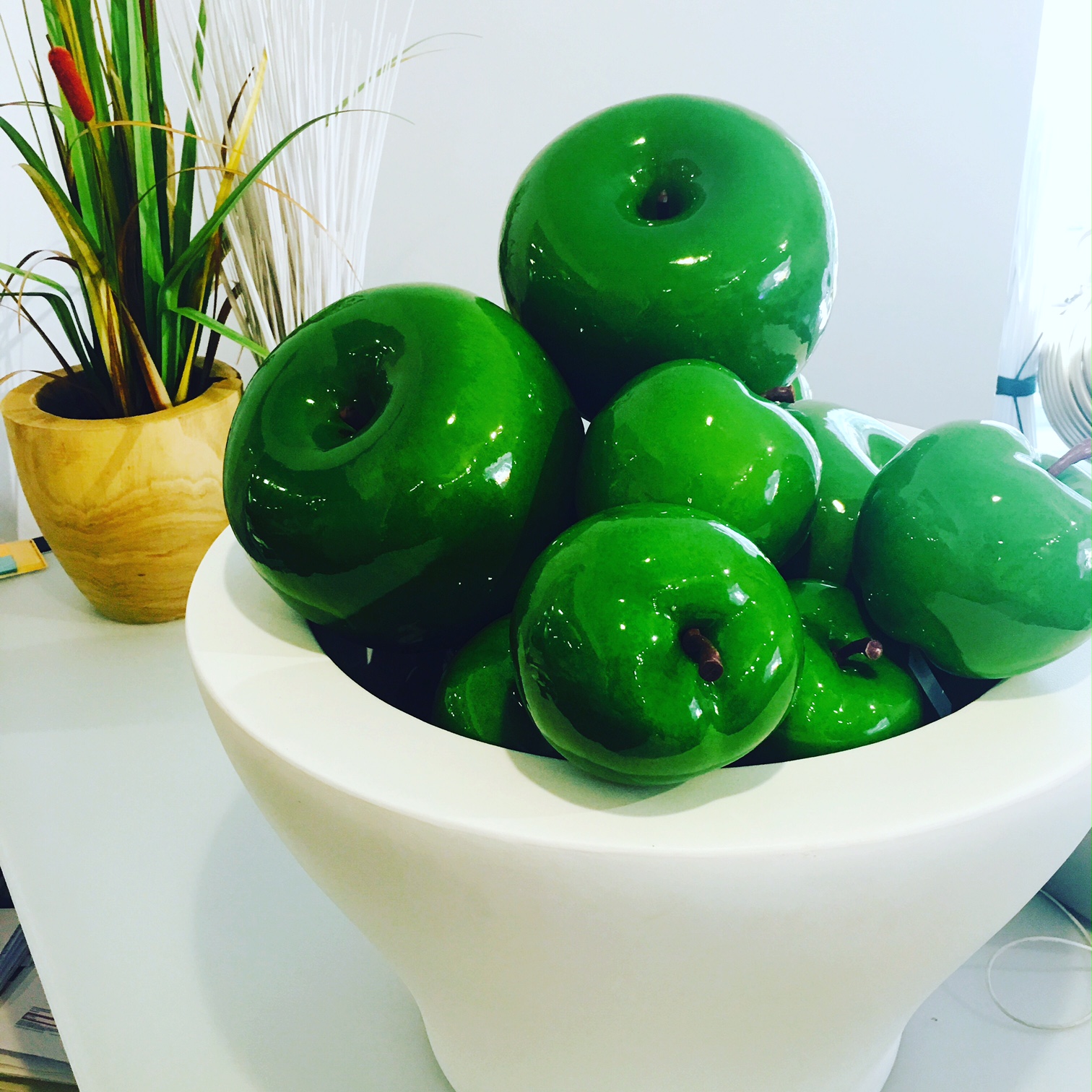 Shiny green decorative apples in a white bowl with a potted plant in the background.