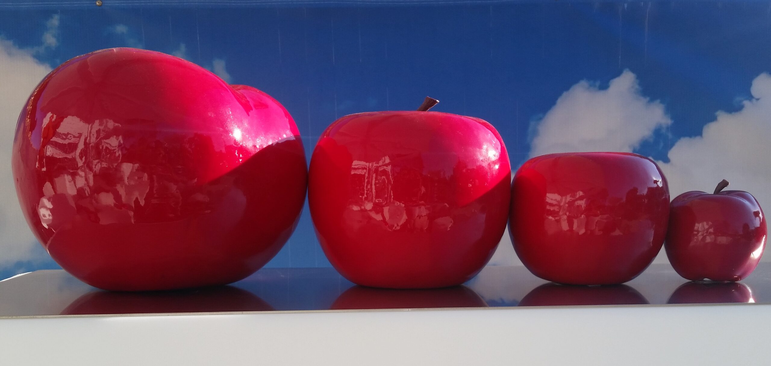 Four glossy red apples of different sizes set against a backdrop of blue sky and clouds