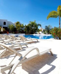 Malta Collection — Sunlit poolside area with rows of lounge chairs, umbrellas, palm trees, and a clear blue sky