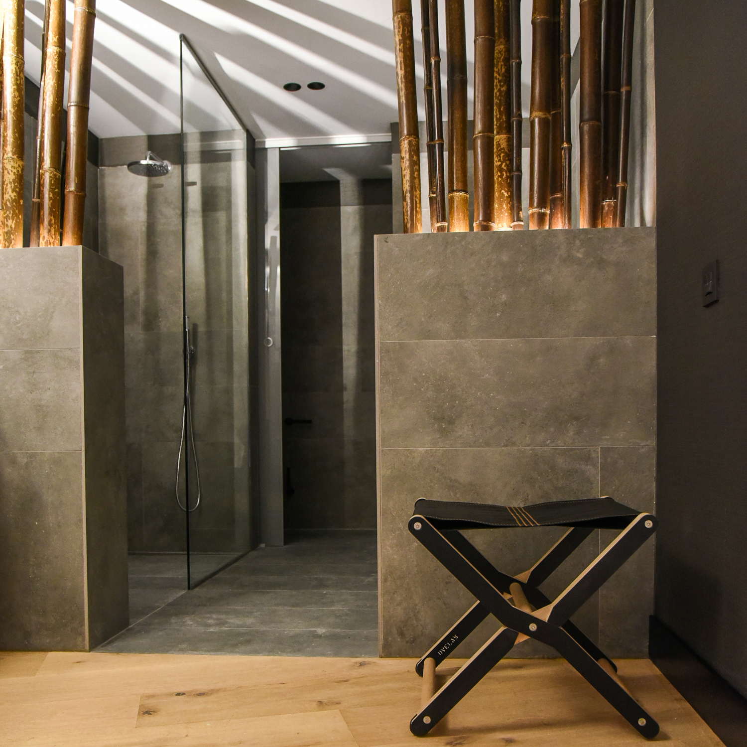 A modern bathroom with gray tiles, a glass shower, & a black folding Stool near bamboo stems.