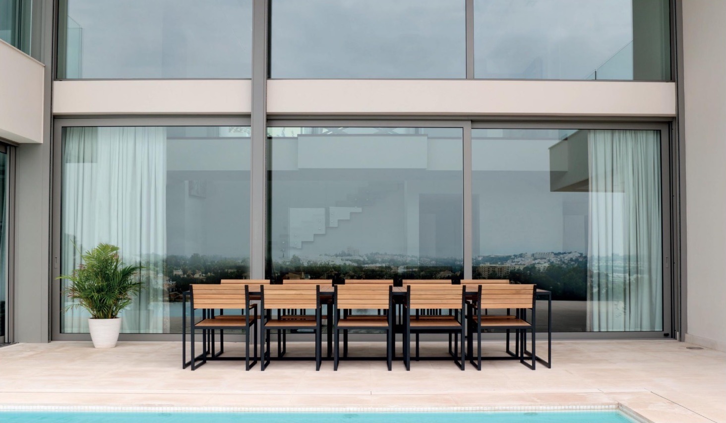 Modern patio with a large glass window, dining table, chairs, and a potted plant by a pool.