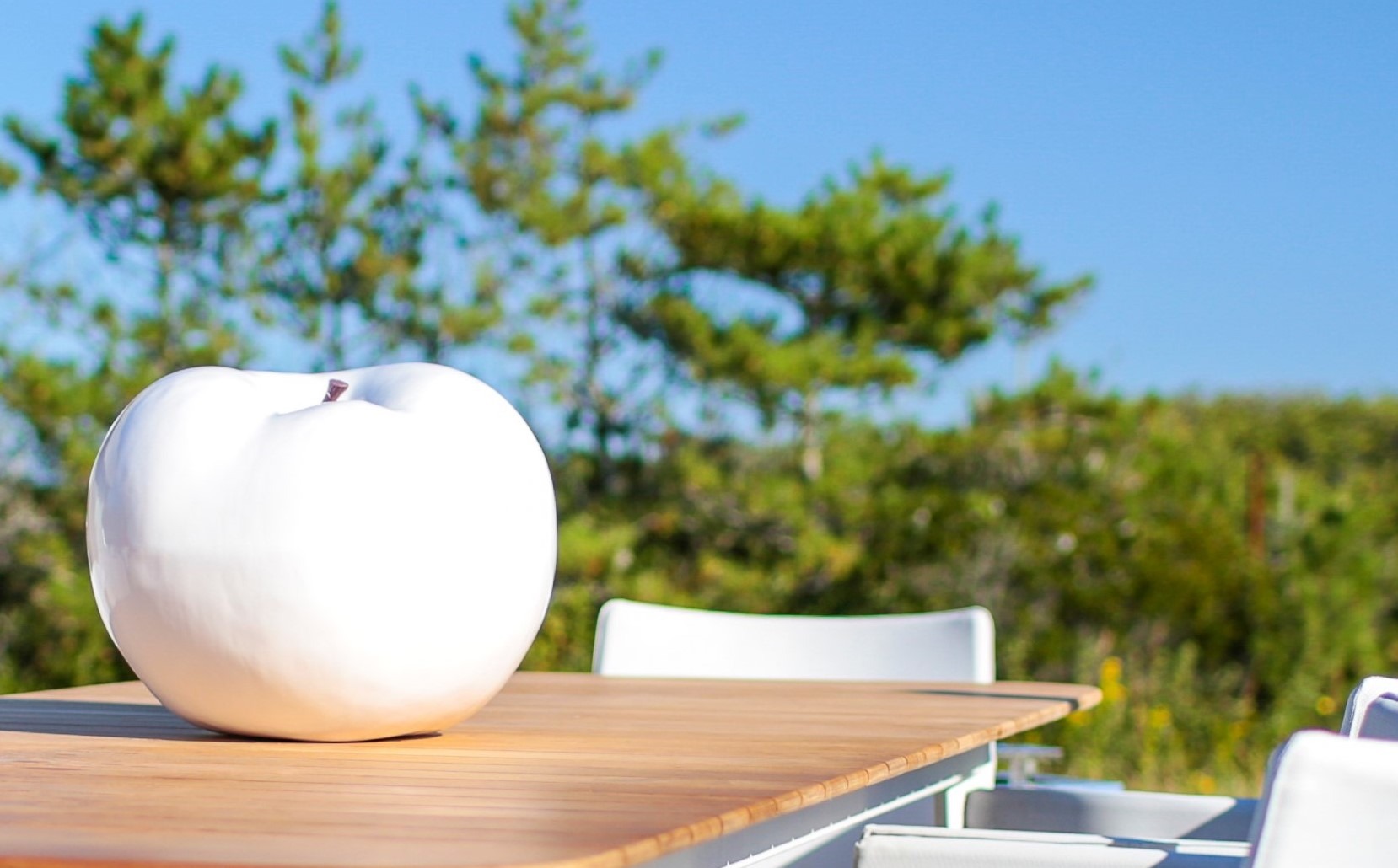 Giant white apple sculpture on an outdoor wooden table with trees in the background.