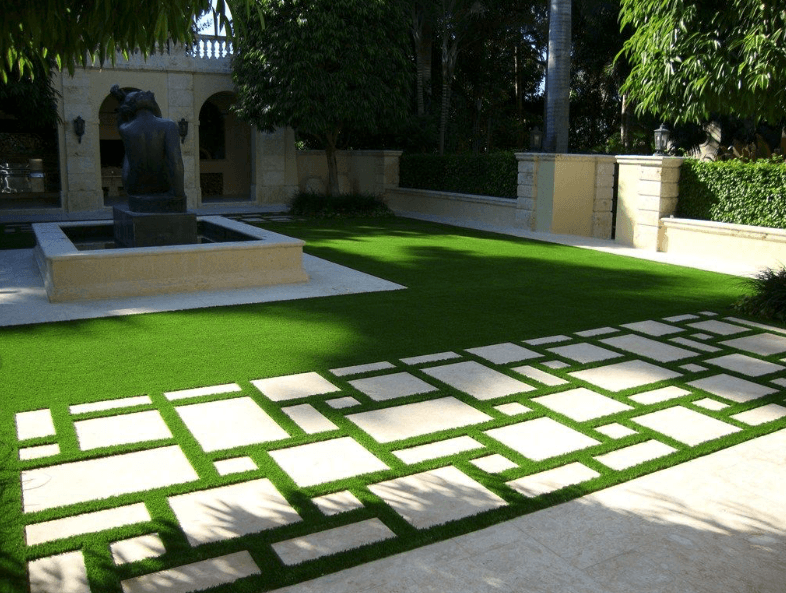 A beautiful garden with artificial grass at its center, surrounded by a geometric pathway, a fountain, and lush trees.