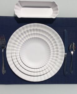 On a blue placemat, there's a white plate with a ridged design, with a silver fork, knife, and spoon, and a white bread plate on top.