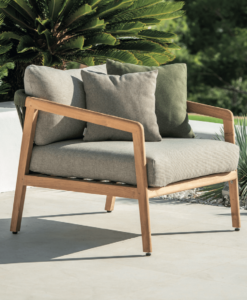 Modern wooden armchair with cushions on an outdoor patio, surrounded by green plants.
